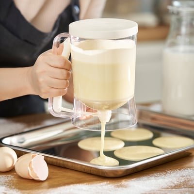 Person pours batter from a clear jug into a muffin tray. The jug has a handle and measurement markings. A bottles and eggshells are nearby.