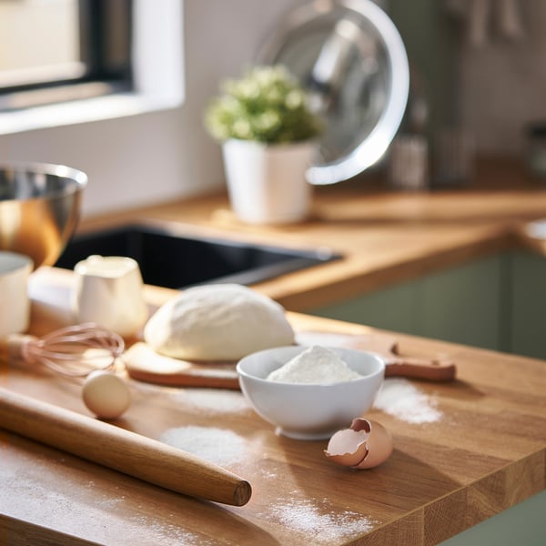 Wooden rolling pin on flour-dusted cutting board with dough, eggshell, bowls, whisk, and potted plant.