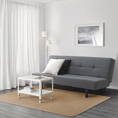 Minimalist living area with grey sofa-bed, white coffee table, and floor lamp against white wall with curtain.