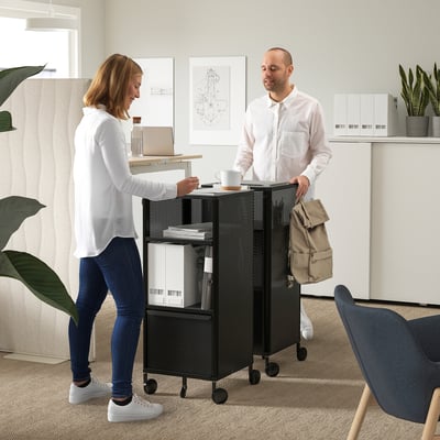 Office scene: two discussing near BEKANT storage with cable management, adjustable shelves, casters.