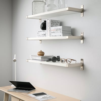 Three white wall shelves, each holding decorative items and books, installed above a light wooden desk with a laptop and papers.