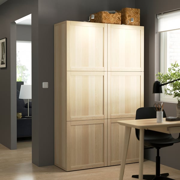 Wooden BESTÅ cabinet in office, between window & black chair. Contrasts dark walls, topped with woven boxes. Left: lamp & plant in adjacent room.
