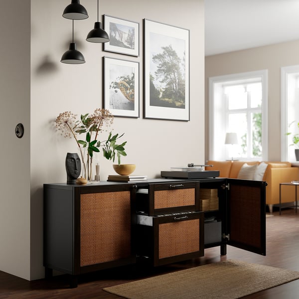 Dark wood sideboard with woven doors, open displaying storage and drawers, above it black pendant lamps and framed art.
