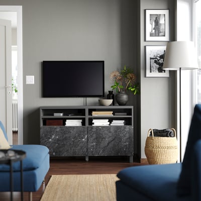 Modern living room with BESTÅ tv stand in black marble and wood, holding a tv, books, and vases. Blue couch and natural basket are visible.