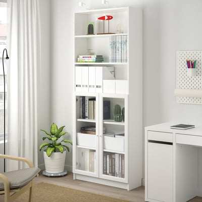 White BILLY bookcase with glass doors, holding books and decorations, beside a wooden chair and a small white desk, with a plant nearby.