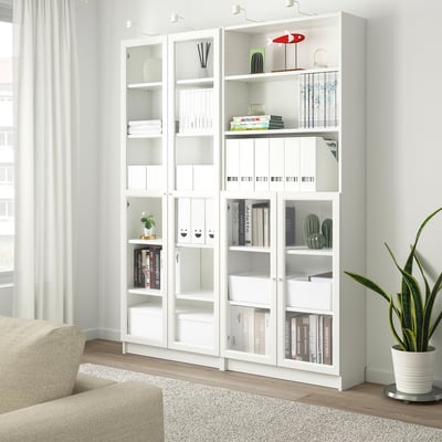 White BILLY bookshelf in living room, glass doors display books, near beige sofa and large window.