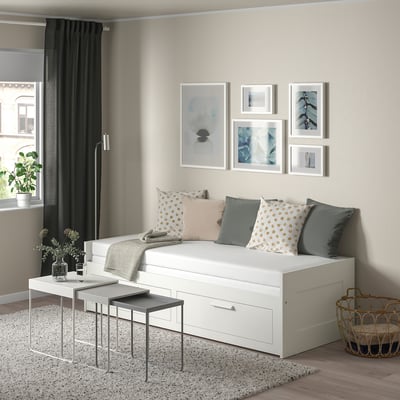 Modern white BRIMNES day-bed with storage and pillows, grey curtains, ample art, grey flooring.