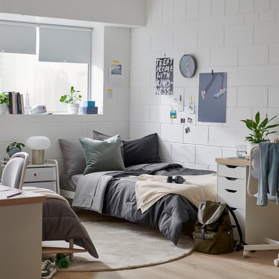 Bedroom with grey comforter, dark green pillows, rug, desk, chair, backpack, posters, clock.