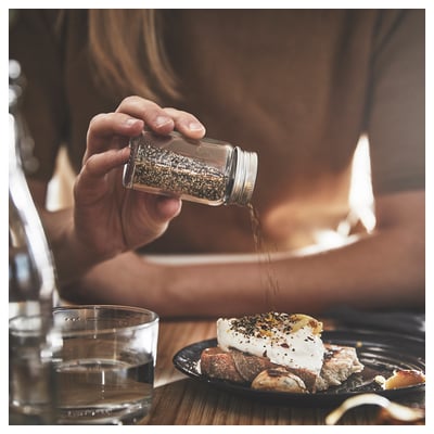Person seasoning food on plate with grinder shaker. Clear glass jar with metal lid, held in hand over dish, light shining through.