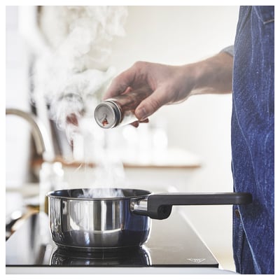 A person in a blue apron grinds pepper in a kitchen, pouring it into a black saucepan on the stove.
