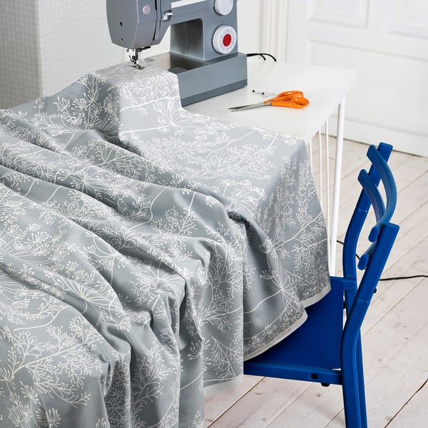 Grey cotton fabric with white floral pattern draped over sewing table, containing sewing machine, scissors, and pins, ready for crafting.