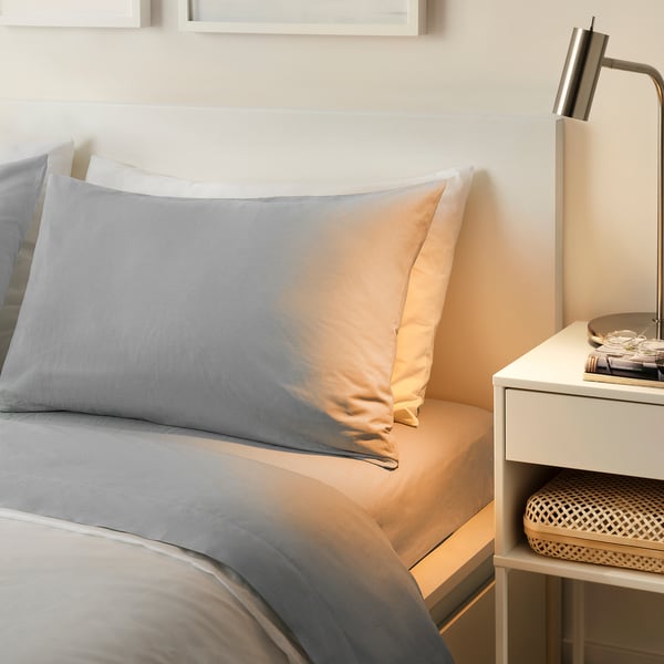 Modern minimalist bedroom with DVALA grey cotton sheet set. Soft fabric, simple design. Large bed next to a small bedside table with lamp.