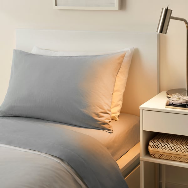 Grey DVALA sheet set on bed, beside table with lamp, books, basket.