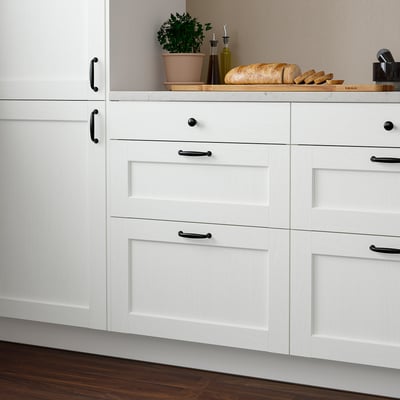 Modern white kitchen cabinets with black handles; small, sleek drawers, larger cabinets to right.