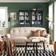 Modern living room with green walls, beige sofa, black coffee table, chequered rug, rocking chair, bookcases, lamp, plant.
