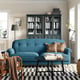 Cosy living room with ESSEBODA blue sofa, dark green bookshelf, black and white rug, wooden chair, floor lamp, and potted plant.
