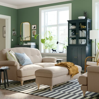 Living room with beige l-shaped sofa, yellow pillows, striped rug, black cabinet, green walls, large window, gold mirror, plants, and wooden chair.