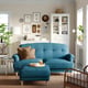 Living room with blue ESSEBODA sofa, brown cushions, matching ottoman, wooden cabinet, wall art, floor lamp, and plants.