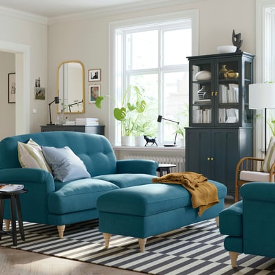 Modern living room with teal ESSEBODA sofa, striped rug, black cabinet, and large window.