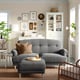 Cosy living room with grey ESSEBODA sofa, wooden legs, brown cushions, white cabinet, green plants, wooden stool, trays, books, and wall art.