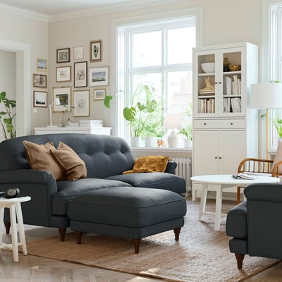 Living room with grey sectional, brown cushions, white furniture, bookshelf, plants, window, art.