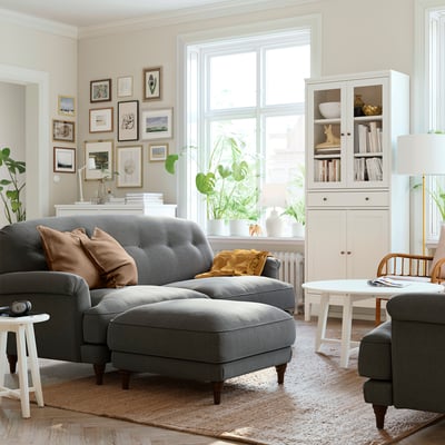 Modern living room: grey sectional sofa with ottoman, brown pillows, white rug, plants, bookshelves, and wall art.
