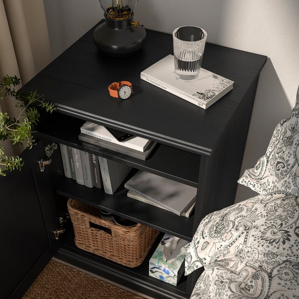 Bedside table with books, glass, plant, and digital clock on top; basket and tissues below.