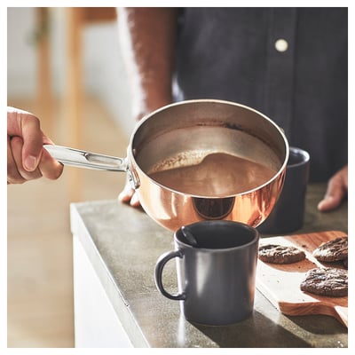 FINMAT copper saucepan, silver handle, dark liquid inside, held by hand, grey mug beside, cookies nearby.