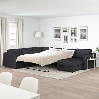 Modern dark grey FINNALA sectional sofa-bed in minimalist living room with photos, plant, lamp, and wooden floor.