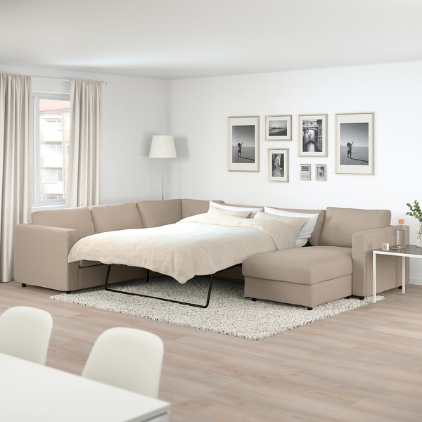 Beige FINNALA sectional sofa-bed with chaise, black & white photo gallery, side table with plants, tall lamp, bright room with rug.
