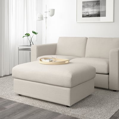 Beige upholstered footstool with storage, tray, and glass, near window with plants and curtains.
