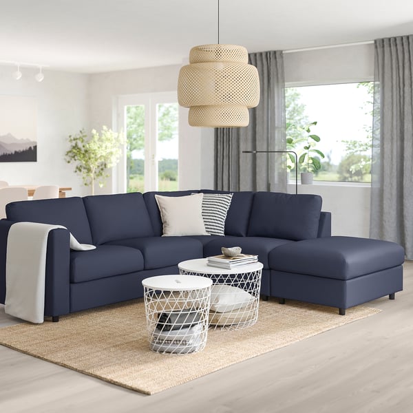 Modern blue sectional sofa with pillows, light wood floor, rug, mesh tables, window with curtains, pendant light.