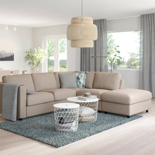 Modern living room with FINNALA beige sectional sofa, grey rug, white coffee table, and large light fixture. Windows let in natural light.