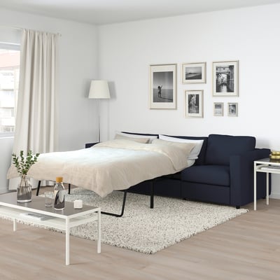 Modern bedroom with FINNALA navy blue sofa-bed, neutral decor, black and white photos, grey rug, white coffee table, and large window with curtains.
