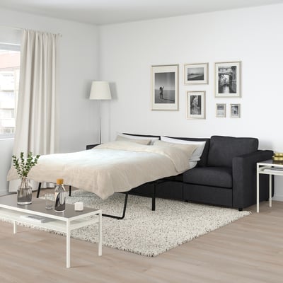 Modern bedroom with FINNALA grey sofa-bed, black frame, by large window, light rug, white coffee table, black bottles, vases, plant, and wall art.