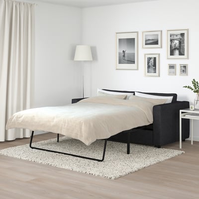 FINNALA black sofa-bed with beige cushions in minimalist bedroom, surrounded by furniture and decor.