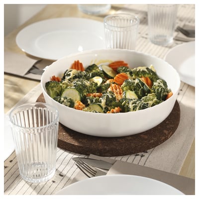 A white bowl of steamed vegetables sits on a wooden table, accompanied by clear glass cups and plates.