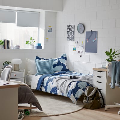 Minimalist bedroom with blue floral bedding, rug, wooden chest, and green backpack. White walls and plants add freshness.