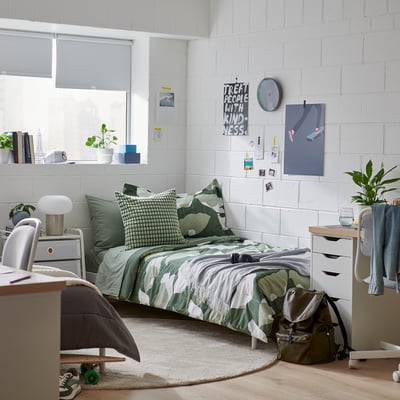 Bedroom with GETRUTA green bedding, size twin, plants, desk, and wall art. Green floral print blanket, white-brick walls. Small, cosy, well-lit.
