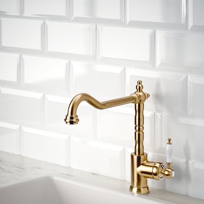 Gold kitchen tap with swivel spout and white handles on white tiled wall.