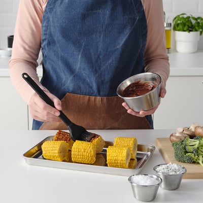 A person in an apron holds a bowl of sauce, brushing corn on a tray with another hand. Beside the tray, small bowls hold dipping sauces.