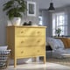 Bright yellow dresser with three drawers, a white vase, and a plant on top. Positioned near a bed with blue curtains.