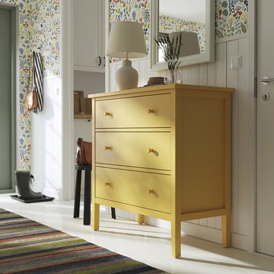 Bright yellow GULLABERG chest, three drawers, wooden handles. Topped with beige lamp and dried branches. Set against wallpapered wall with mirror.