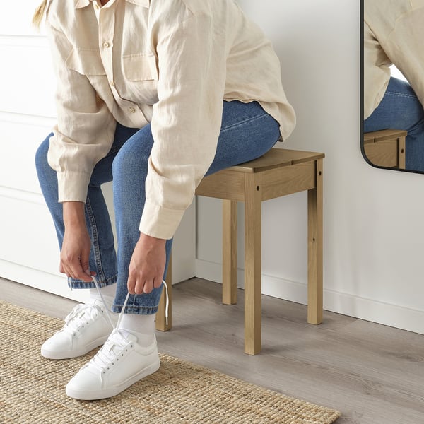Person sitting on wooden stool, tying white trainer, light background.