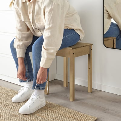 Person sitting on wooden stool, tying white trainer, light background.