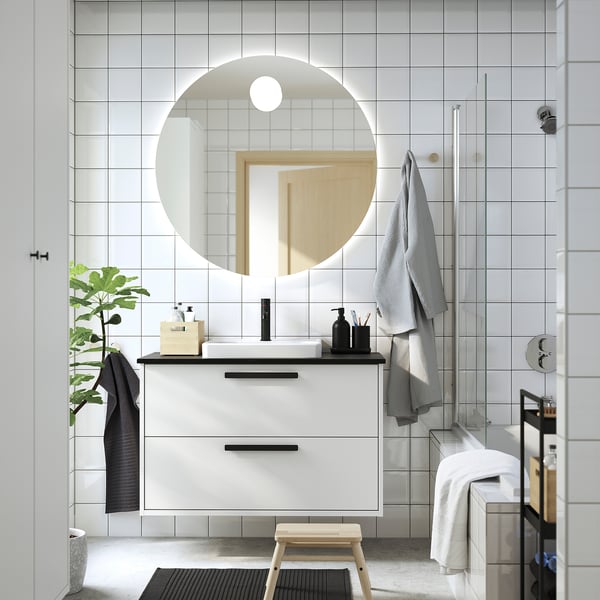 Modern bathroom with white cabinet, black accents, round mirror, grey towels, wooden stool, plant, and wall storage.