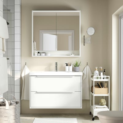 Modern white bathroom vanity with 2 drawers, mirror cabinet, small plant, toiletries.