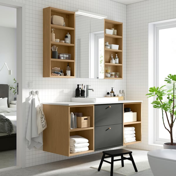 Modern bathroom featuring HAVBÄCK light wood vanity, dark grey drawers, and ceramic basin. Shelves hold toiletries, towels below, and plants beside.