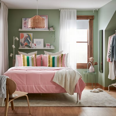 A bright green bedroom with pink pillows, colourful art, and a woven pendant light.