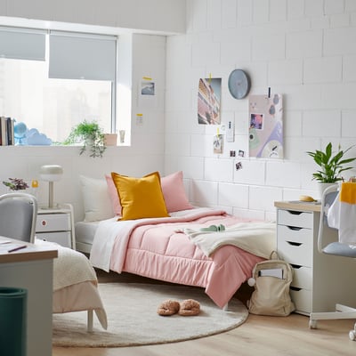 Cosy bedroom with twin beds, white brick walls, and a large window. Pink & yellow pillows, soft rug, and plants decorate the room.